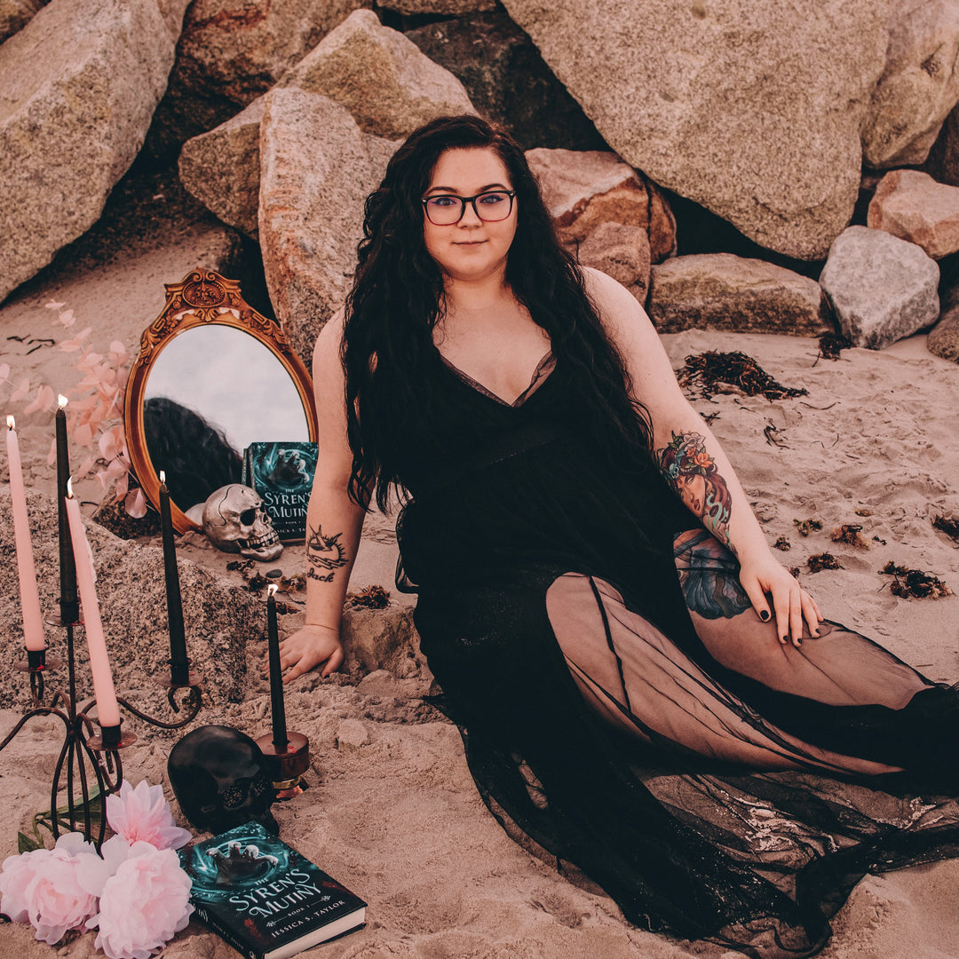 Image of Jessica S. Taylor; a woman with long black curly hair in a black gauzy dress sitting on a beach. Background is rocks, sand, and the borders of the image are filled with a candelabra, flowers, a mirror, skulls, and copies of Jessica's books.