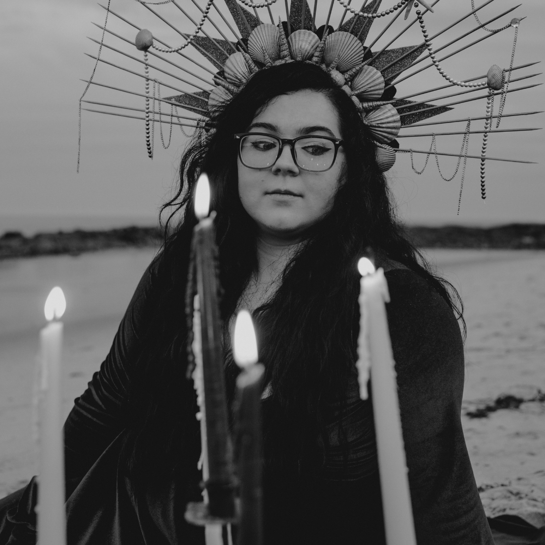 Black and white image of a woman with long wavy dark hair and glasses looking out over a set of 4 candles. She is wearing a long sleeve velvet dress and a crown with shells and pearls, and is sitting on a beach.